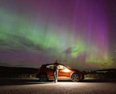 The Northern Lights in the north coast of Scotland. There are green and purple ripples across the sky. Gary Macleod stands in front of his car under the lights.