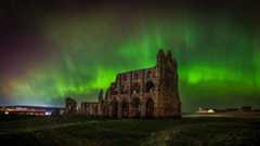 The Northern Lights in green and edged with red over Whitby Abbey in North Yorkshire. The brightest parts look like ribbons in the sky. The ruins of the abbey stand underneath with fields around them. there is a house lit up in the background.