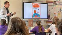 Tom stands in front of a smart board with children in purple school jumpers watching him describe how to draw the characters from his book.