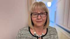 Ildiko Veres. She has a blonde bob with a fringe, blue eyes, wearing black glasses, a grey tweed jacket and silver necklace.