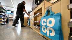 A blue bag-for-life with a big white 'Co-op' logo is seen hanging on a peg next to the checkout in a Co-op store with a worker in the background stacking shelves, in London in 2021.