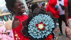 A woman in a market in Kenya holding a bouquet made out of blue coloured Kenyan shilling banknotes. Behind her is a stall selling teddy bears.
