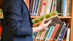 A child wearing a navy blue school blazer looks through a history book in a library. They are stood in front of a bookshelf which has dozens of other history books on it.
