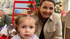 A woman with tied up brown hair is crouched down behind a child, smiling at the camera. She is wearing a dark top and a light brown jacket. Behind her is a Christmas tree. The child has fair hair, tied into pig tails, and is wearing a blue top and multi-coloured bib.