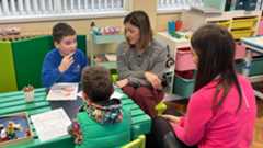 Two women with brown hair sit at a small green table in a school, speaking with two children. The boys both have short dark hair and have colouring materials in front of them. One is wearing a blue sweatshirt and the other is in a green sleeveless jacket with a patterned hoodie underneath.