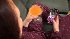 Woman sat on a sofa holding a cocktail in one hand and her phone in the other. She is watching a live stream from a bar in Manchester. (Stream image has been added to phone by Photoshop for illustration purposes)