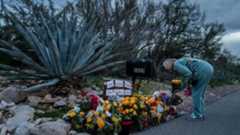 A woman in a teal sweatsuit bends over and lays flowers at a memorial for Nancy Guthrie