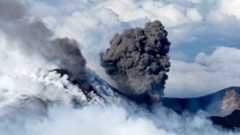 A black plume erupts from Mt Etna with clouds behind it.