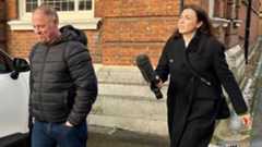 Raymond McDonald, a man in his fifties, is walking away from Philippa Goymer who is a BBC journalist. She is dressed in black and has long dark hair and is carrying a large microphone. He has a black jacket, blue jeans and has his hands in his pockets. Behind them both is a brick built building and a white car is parked to the side.