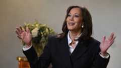 A photograoh of Kamala Harris waving her hands in the air. She is wearing a dark coloured suit with a white shirt.