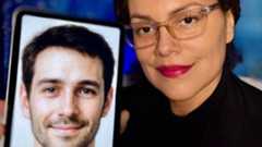 A woman with clear glasses and bright red lipstick smiles as she holds her phone to the camera showing a picture of her AI companion. On the phone is a picture of a man with short cropped hair, a slight beard and smiling eyes.