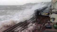 A damaged section of the coastal railway at Dawlish in 2014, with waves washing over broken track where the sea wall has collapsed. Houses sit close to the line on the right under a grey sky.