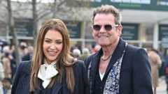 A woman with long-brown hair and wearing a dark pinstriped suit with a white scarf stands next to a man with a dark coloured suit and blue and white patterned scarf. He has short grey hair which is swept back and spiked up. He has a chain necklace and sunglasses. They are both smiling.