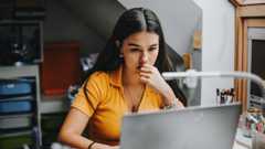 A young woman in an orange top looks at her laptop while chewing her finger