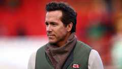 Headshot of Ryan Reynolds at the football pitch. He has dark brown hair and is wearing a white jumper with a green gilet over the top, There's a brown scarf-like item around his neck.