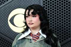 Lola Young has long black hair, she is wearing a dark green jumper with a large white collar over the top, she has a orange and blue striped tie on