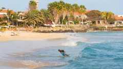 A man surfing near the coastline of Santa Maria, Sal Island, Cape Verde. The water is a pale blue and he is riding a small wave. There are palm trees and white buildings with terracotta-coloured roofs in the background.