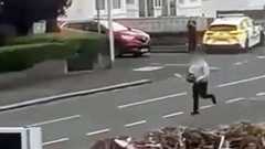 Grainy footage capturing a man wearing black trousers and a grey top running down a residential street while brandishing a chainsaw. A parked police car and a lone officer can be seen in the background