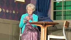 A woman sits on a chair at a small table on stage. She has grey hair and glasses, and is holding a needle and some material