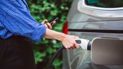 A hand holding an EV charger at a silver car. The person is wearing a striped blue and white shirt, and black trousers