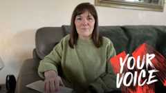A woman with shoulder-length dark hair, wearing a green jumper, sits on a sofa looking at the camera