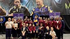 A group of school children standing infront of darts boards and some adults are also in the photo. They are smiling and some have their thumbs up. Their school uniform is maroon with a yellow crest.