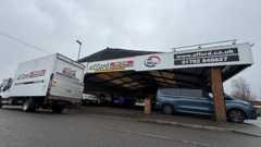 A garage with vehicles parked under a large roof. A white van is parked at the side of the road in front of it.