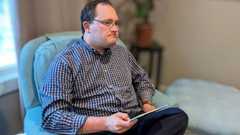 Nicholas Strahl, wearing glasses and a checked shirt, sits in an armchair holding a tablet.
