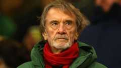 Jim Ratcliffe, with a grey stubbly beard, watches football in the stands wearing a red scarf and green jacket