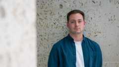 Ben is a 25-year-old man and stood against a pale white stone background outside. He is in a blue shirt with a white t-shirt underneath, has brown hair combed to the side and has one hand in his pocket