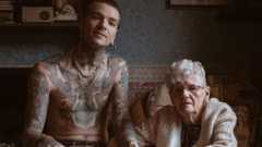 The photo Tom took, one of the winning photos in Portrait of Britain. Adam and his nan Janet sit next to each other in her house, with Janet putting both hands around Adam's left hand. He is topless, revealing tattoos of mythological creatures that cover his torso and arms. Adam has gold hoop earrings and a gold necklace. He wears brown trousers and converse-style shoes. Janet wears maroon leather shoes, brown trousers and a thick cream cardigan. She has short silver hair and wears glasses.