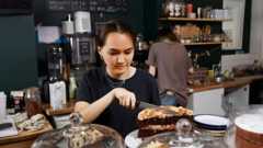 A young woman in a cafe cutting a slice of cake with a neutral expression on her face