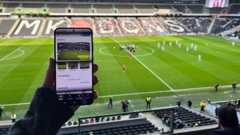 Image of someone sitting in a football stadium holding up a mobile phone, with images of a football pitch on it. In the background can be seen a number of empty stadium seats, a football pitch, football players and officials.