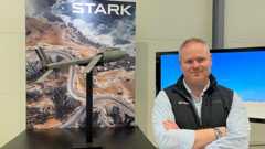 Mike Armstrong - managing director of drone company Stark - looks at the camera, arms folded, wearing blue shirt and dark blue branded gilet. Next to him is a model of a drone and the Stark logo on a board with a drone shot of a coastal area.
