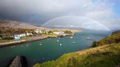There is a sea loch with brightly coloured boats and a rainbow might looks like a bridge connecting the land on either side. There is a row of houses and a pier on one side of the Loch