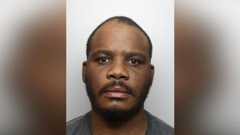 Kennedy Jimmy is looking at the camera in a police custody shot. He has very short black hair and a black beard and moustache. He is wearing a green T-shirt.