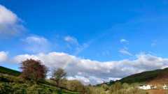 an undulating valley with trees, white houses, low cloud and a rainbow under blue sky