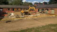 An old red brick stable block, with a digger in front of, where building work is underway. There is a large area of gravel in front with some grass closer to the front.