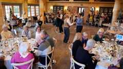 Groups of older people sit around round dining tables as they enjoy afternoon tea in a large dining room as people swing dance in the centre of the room