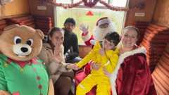 Jessica with her son Rory is sitting in a carriage with Santa, a woman wearing a Santa cloak and a life-size teddy bear. Father Christmas and Ms Lymn are waving at the camera. They are sitting in an enclosed carriage decorated with tinsel.