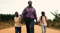 Actor Sope Dirisu walks down a dusty road whole holding the hands of young actors Godwin Chiemerie Egbo and Chibuike Marvellous Egbo. Sope is wearing a purple striped shirt and jeans. The boys are wearing jeans and T-shirts.