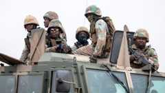 A group of six soldiers wearing camouflage uniforms and helmets riding on top of a large armored military vehicle. Some of them are holding rifles, and a few wear goggles or face coverings.