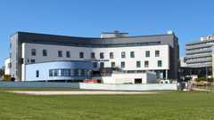 The hospital is a large, four storey white and grey building, part of the building is blue and extends from the front of the hospital.
