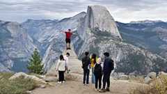 People take photos from Glacier Point with Half Dome in the background at Yosemite National Park. A man in a red shirt is standing at the edge of a popular photo spot with is arms spread out.