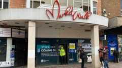 The outside of the former Market Walk shopping centre, with a man in a high-vis jacket in the doorway, and three other people to the right. A man is by a cash machine to the right. There is a white frame round the building the front boarded up with signs on it.