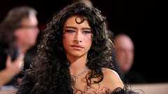 Odessa has brunette hair, she's posing for a picture on a red carpet, she has a nose ring and a silver necklace on. In the background there are blurred photographers.