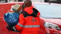 A Royal Mail postman holding two large parcels. He is standing in front of a red Royal Mail van