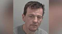 A mugshot of a man in a grey T-shirt, against a grey backdrop. He has short brown hair and greying sparse beard. His eyes are partially closed.