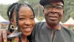 Ifunanya Nwangene, with braids each decorated with a shell and wearing large flower-like earrings, and her father Christopher, who has his arm around her shoulder with a phone in his hand. They are both smiling at the camera.