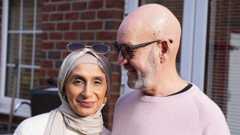 This photograph is of Sahir and Dennis in their garden. Sahir, on the left, is wearing a creamy jumper with a soft cream headscarf. She has a pair of glasses on her head and is wearing gold necklaces. Dennis has his arm around Sahir and is looking at her as she smiles at the camera. He's wearing a light pink/lilac knit jumper with a black t-shirt underneath. He's wearing glasses and has a beard.